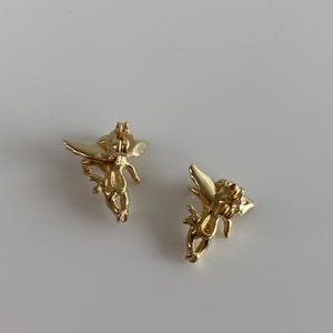 May include: Two gold-colored angel earrings. Each earring features a detailed depiction of an angel with outstretched wings. The earrings are set against a plain white background, highlighting their intricate design and metallic sheen.