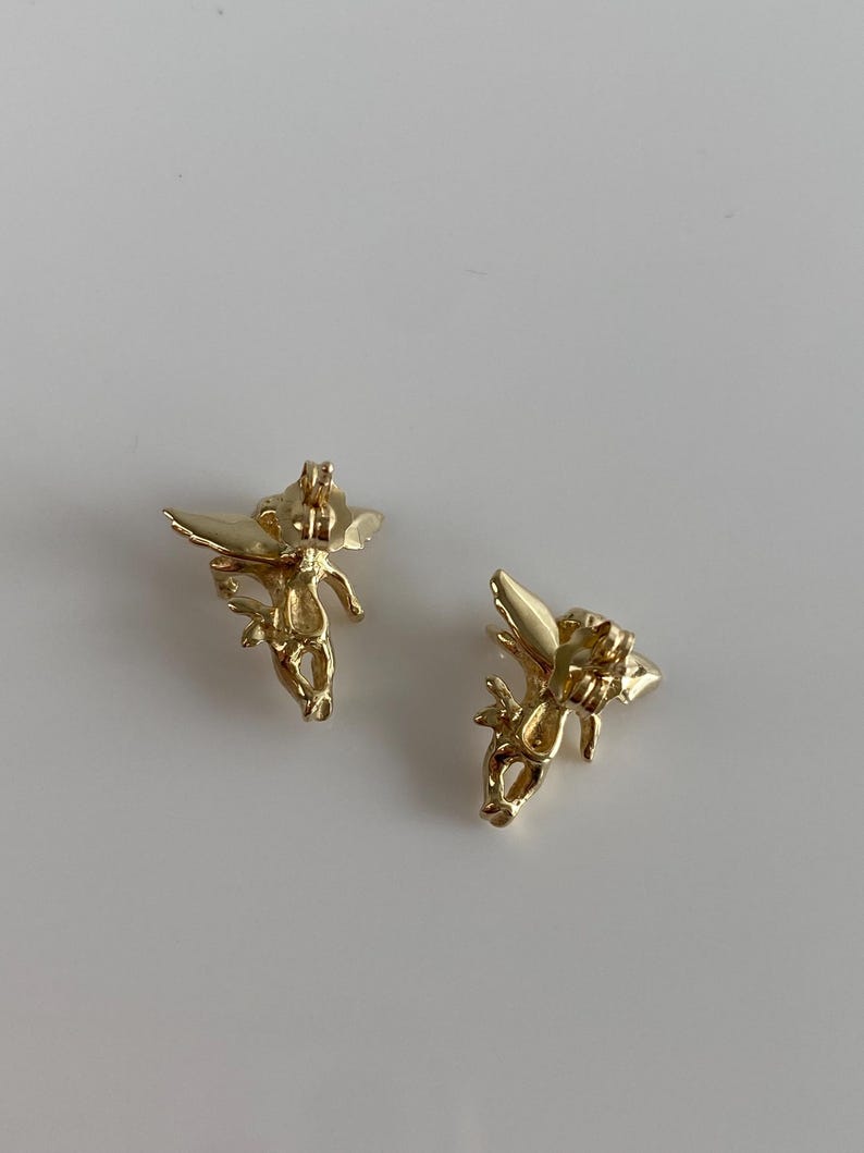 May include: Two gold-colored angel earrings. Each earring features a detailed depiction of an angel with outstretched wings. The earrings are set against a plain white background, highlighting their intricate design and metallic sheen.