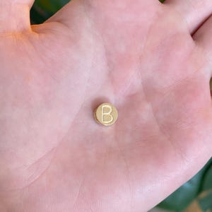 May include: A small, circular, gold-colored metal bead featuring the capital letter B embossed on its flat surface. The bead has a smooth, polished finish and a cylindrical shape with a hole running through the center for threading onto a cord.