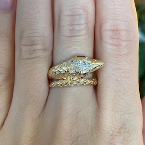 May include: A gold-colored snake ring with a textured design. The snake's head features small, clear gemstones. The ring wraps around the finger, creating a unique and eye-catching accessory. The ring is worn on a hand.
