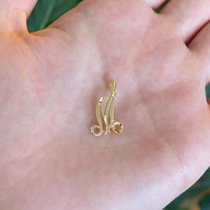 May include: A small, polished gold pendant shaped like the cursive letter M. The pendant features a reflective, metallic surface with a pointed bail at the top for attachment to a chain. The design has flowing, elegant lines and a bright yellow gold tone.