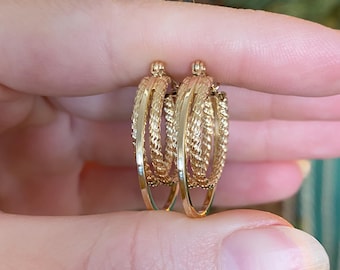 May include: A pair of gold hoop earrings with a textured, braided design. The earrings are held between fingers, showcasing their intricate details and warm, golden color. The hoops have a layered appearance, adding depth to the design.