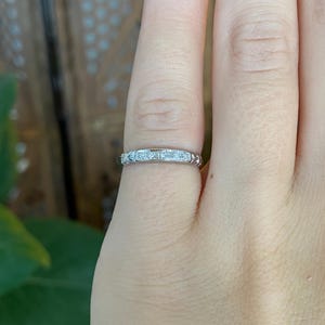 May include: A silver-toned metal band ring featuring a series of small, clear, square-cut stones set in a channel along the center. The band has a polished finish and decorative engraved detailing on the sides of the metal setting.