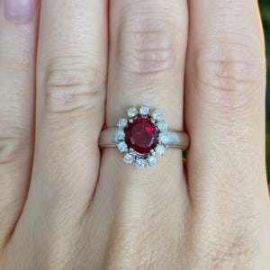 May include: A silver-toned ring featuring a central oval-cut deep red gemstone surrounded by a halo of twelve small, round, clear white stones. The band is smooth and metallic, providing a clean contrast to the vibrant red center stone.