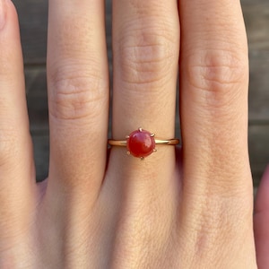 May include: A gold ring with a red gemstone set in a prong setting. The ring is on a person's finger.