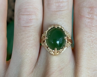 May include: A gold-toned ring featuring a large, oval, deep green gemstone. The ring has an ornate, filigree-style setting with intricate details around the gemstone. The ring is worn on a finger.