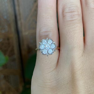 May include: A silver-toned ring featuring a cluster of seven round, clear, faceted stones arranged in a floral pattern. The central stone is surrounded by six identical stones, all set in a metal frame with small prongs securing each individual gem.