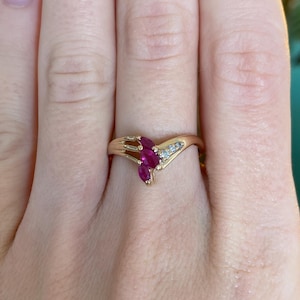 May include: A gold ring with three marquise-cut ruby gemstones and two small diamonds. The ring has a delicate, curved design, with the gemstones set in a cluster. The ring is worn on a finger.