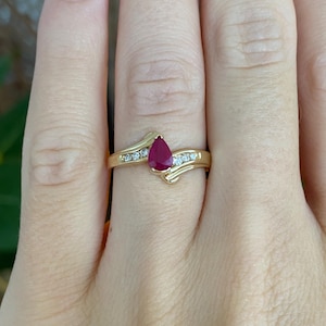 May include: A gold ring featuring a central pear-shaped red ruby gemstone. The band has a bypass design with three small round white diamonds set into each side of the shank, flanking the ruby in a symmetrical arrangement on the polished gold band.