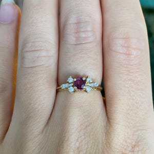 May include: A gold ring featuring a central round-cut deep pink gemstone flanked by four small round-cut clear stones and two baguette-cut clear stones arranged in a cluster setting on a thin, polished gold band.
