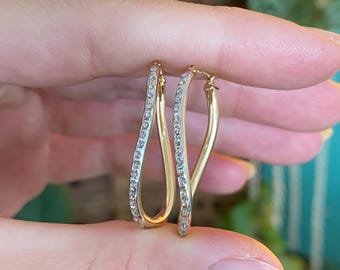 May include: A pair of gold-toned hoop earrings. The earrings have a curved, elongated oval shape. One side of each earring is encrusted with small, clear gemstones. The earrings are held between fingers against a blurred background.