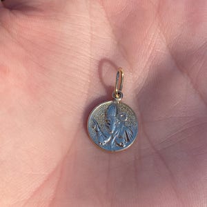 May include: A gold pendant featuring a detailed image of a saint. The pendant is round and has a textured surface.