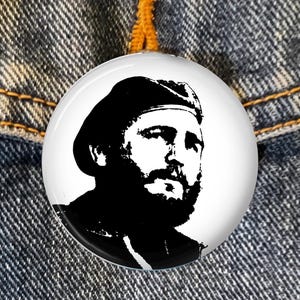 May include: A round, white button featuring a black and white portrait of a man with a beard and beret. The button is set against a denim jacket background.