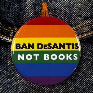 Puede incluir: Un botón con los colores del arcoíris con el texto "BAN DESANTIS NOT BOOKS" sobre un fondo de mezclilla.