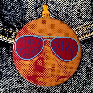May include: A round orange button with a red and blue graphic of a face wearing sunglasses. The sunglasses have the word "WAVES" written in blue on the lenses.