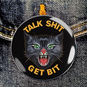 May include: A black button with the text "TALK SHIT GET BIT" in yellow. The button features a black cat with green eyes and an open mouth, revealing its teeth. The button is set against a denim background.