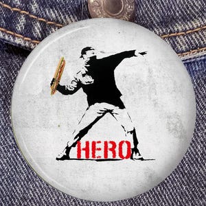 Puede incluir: Un botón redondo con un gráfico de una persona lanzando un sándwich, en un estilo de plantilla en blanco y negro. La palabra "HERO" está impresa en rojo debajo de la figura. El botón está sobre un fondo de mezclilla.