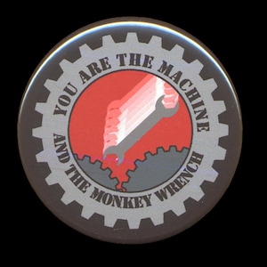 May include: A black and silver button with a red background and a wrench in the shape of a hand. The text on the button reads "You are the machine and the monkey wrench."