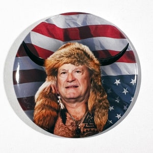 May include: A button featuring a man with a long brown beard and a fur hat, wearing a necklace with a symbol. He is in front of an American flag with a bull's horns.