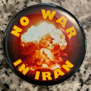 May include: Round button with a black border and a graphic of a nuclear explosion. The words "NO WAR IN IRAN" are in yellow, arched around the image.