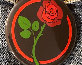 Puede incluir: Un botón negro con un círculo rojo alrededor de una rosa verde con un centro rojo. La rosa está sobre un fondo negro.