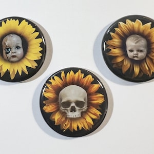 May include: Three round black buttons with a sunflower design. Each sunflower has a different face in the center: a doll, a skull, and a baby.
