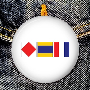 Könnte beinhalten: Ein weißer Knopf mit drei nautischen Signalflaggen. Die Flaggen sind rot mit einer weißen Raute, gelb mit einem blauen Streifen und rot-weiß mit einem blauen Streifen.