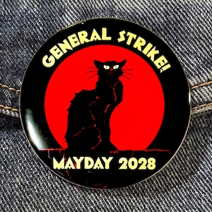 May include: A black and red button with a black cat silhouette in front of a red sun. The button reads "GENERAL STRIKE! MAYDAY 2028".