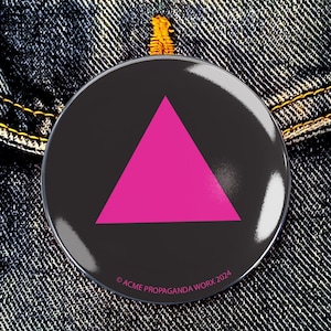 Può includere: Un bottone rotondo nero con un triangolo rosa acceso al centro. Il bottone ha una finitura lucida ed è posto su uno sfondo di denim. Il testo "© ACME PROPAGANDA WORK 2024" è stampato in basso.