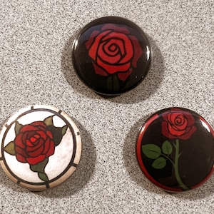May include: Three round pinback buttons with black backgrounds and red roses. The first button features a red rose with a black outline. The second button features a red rose with a white outline and a white background. The third button features a red rose with a green stem and leaves.