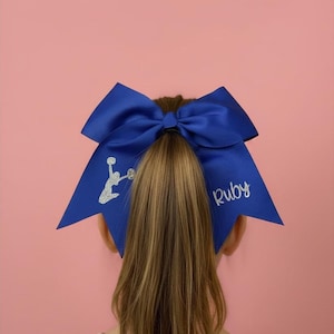 May include: A large royal blue hair bow featuring a silver glitter graphic of a cheerleader in a jump pose on the left side and the name Ruby in silver glitter script on the right side, attached to a high ponytail with a centre knot.