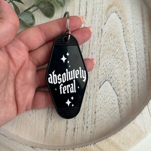 May include: A black motel-style keyring with a diamond shape and rounded edges. The keyring features white text that reads absolutely feral, accented by small white star symbols above and below the text. A silver metal ring is attached at the top.