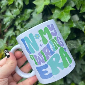 May include: White ceramic mug featuring the phrase "IN MY TWIRLING ERA" in a retro font. The text is in shades of blue, green, and purple, with flower and star details. The mug has a white handle.