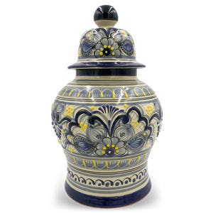 May include: A ceramic jar with a lid, featuring a detailed floral design in shades of blue, gray, and yellow. The jar has a bulbous shape with a matching lid and a black knob. The design includes intricate patterns and decorative elements.