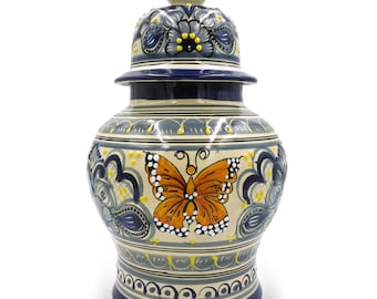 Può includere: Un vaso in ceramica con coperchio, con un design a farfalla dipinto a mano. Il vaso è decorato con motivi floreali blu, gialli e grigi. La farfalla è arancione con punti bianchi. È un oggetto decorativo.