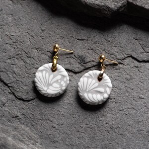 May include: A pair of gray ceramic earrings with a white floral design. The earrings have gold-toned hooks.