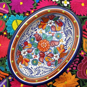 May include: Oval ceramic serving dish with a colorful floral design. The dish has a blue rim with green and brown accents. The floral design features red, orange, yellow, green, and white flowers.