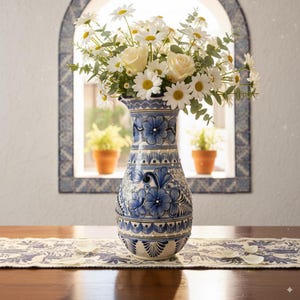 May include: A tall, blue and white ceramic vase filled with white roses and daisies. The vase has a detailed floral pattern and sits on a table with a patterned runner. The background includes a window with potted plants.
