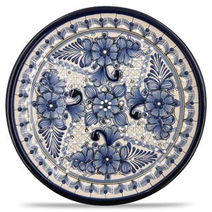 May include: Round ceramic plate with a hand-painted floral design. The plate features a cream-colored center with blue flowers and foliage. The rim is dark blue, and the overall design is symmetrical and detailed, suitable for serving or display.