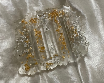Puede incluir: Jabonera de resina transparente con diseño rectangular y ranuras verticales. La jabonera está adornada con copos de pan de oro y plata, creando un aspecto decorativo y elegante. Los bordes tienen una forma ondulada e irregular.