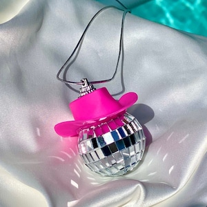 May include: A silver disco ball ornament with a pink cowboy hat charm hanging from a silver chain. The ornament is on a white satin fabric background.