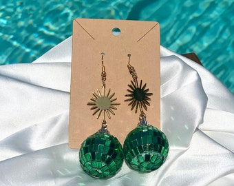 May include: A pair of green disco ball earrings with gold star accents. The earrings are hanging from a cardboard backing.