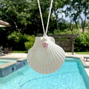 May include: A white seashell with a pink flower charm hanging from a white string. The seashell is hanging over a blue swimming pool.