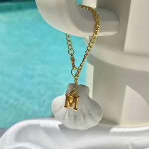 May include: A gold chain necklace with a white seashell charm featuring the letter "M" hanging from a white object.