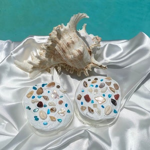 May include: Two white round coasters with a variety of seashells embedded in the surface. The coasters are on a white satin fabric with a large seashell in the background.