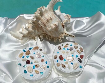 May include: Two white round coasters with a variety of seashells embedded in the surface. The coasters are on a white satin fabric with a large seashell in the background.