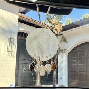 May include: White round car air freshener with a string hanging from the top. The air freshener has five gold rings with different seashells hanging from them. The seashells are white, brown, and black.