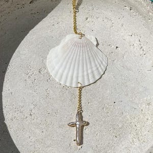 May include: A white seashell pendant with a gold chain and a gold cross charm. The pendant is displayed on a textured, light-colored surface, with a blurred blue water background.