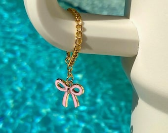 May include: A gold chain with a pink bow charm hanging from it. The chain is hanging from a white surface.