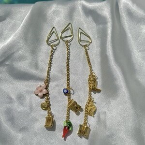 May include: Three gold chains with charms, each featuring a different design. The first chain has a pink flower, a smiley face, and the letter "D". The second chain has a blue evil eye charm, the letter "R", a red chili pepper, and the letter "M". The third chain has a gold angel wing charm, a gold shell charm, and a gold feather charm.